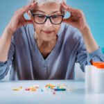 Idősebb szemüveges nő kék ingben ül az asztalnál könyökölve gondterhelten, előtte egy kupac tabletta és kapszula, kék háttérrel.