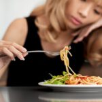 Fiatal szőke lány villával piszkálja a tányérban lévő spagettit.