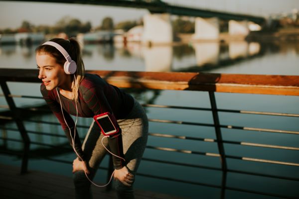 Fiatal nő sportöltözetben és fejhallgatóval a fején pihen futás után egy hídon, mögötte folyó.