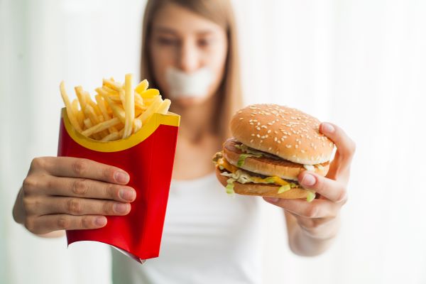 Fiatal nő leragasztott szájjal egyik kezében gyorséttermi sült krumpli, másikban hamburger.