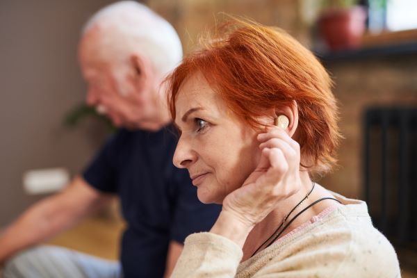 Vörös hajú idős fülében hallókészülékkel, a háttérben idős férfi ül.