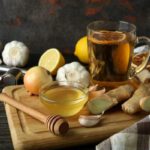 Vágódeszkán egy pohár tea, kis üvegtálban méz, gyömbérdarabok, fokhagyma, hagyma, citrom, narancskarika.