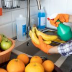 Kockás inges nő a mosogató fölött egy köteg banánt tart, amelyre másik kezével ráfúj egy szórófejes flakonból, körülötte narancsok, almák.