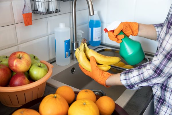 Kockás inges nő a mosogató fölött egy köteg banánt tart, amelyre másik kezével ráfúj egy szórófejes flakonból, körülötte narancsok, almák.
