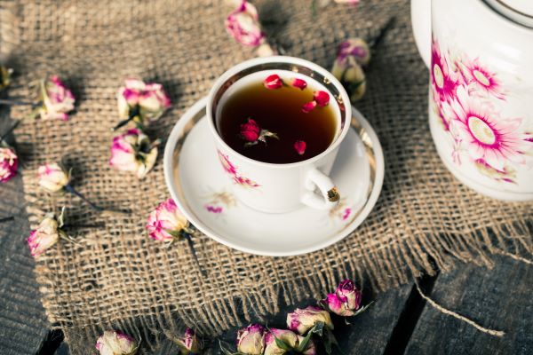Virágos csészében tea rózsaszirmokkal, mellette virágos teáskanna, körülötte elszórva rózsabimbók.
