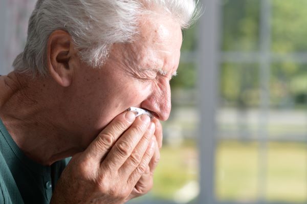 Idős férfi összeszorított szemmel zsebkendőt szorít a szájához.