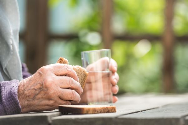 Közeli fotó idős nő kezeiről, amelyekben egy szelet kenyeret és egy pohár vizet tart.