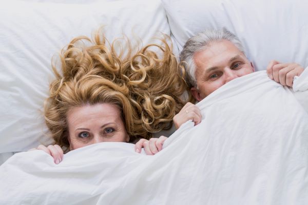 Középkorú pár fekszik az ágyban a paplan alatt, csak a fél fejük látszik ki.