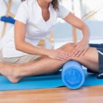 Fehér köpenyes fizioterapeuta nő foglalkozik a kék matracon fekvő férfi lábával.