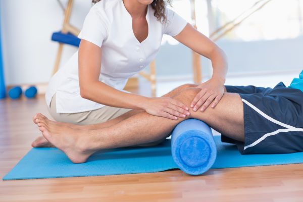 Fehér köpenyes fizioterapeuta nő foglalkozik a kék matracon fekvő férfi lábával.
