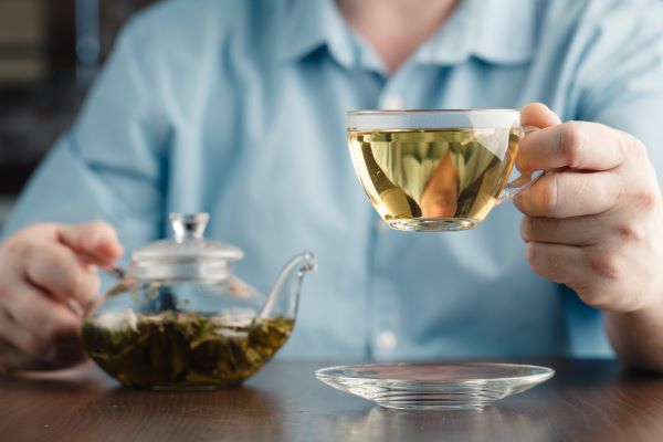 Kék inges férfi egyik kezével teáskannát fog, másikkal üvegcsészét zöld teával.
