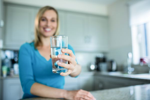 Szőke nő a konyhában egy pohár vizet tart a kezében.