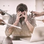 Férfi ül a laptopja előtt gondterhelten, míg kezek két oldalról papírt, mobiltelefont, ceruzát, tabletet nyújtanak felé.