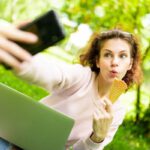 Fiatal nő a parkban laptoppal az ölében és fagylalttal a kezében szelfit készít a telefonjával.