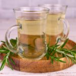 Gyógyítás, fogyás és stresszcsökkentés rozmaringteával Fatálcán két pohár rozmaringtea, mellette rozmaringágak.