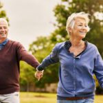 Idősebb nő és férfi kézen fogva vidáman mennek egy parkban.