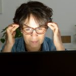Középkorú szemüveges nő halántékát fogva ül a monitor előtt.