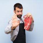 Szakállas férfi nagy darab nyers húst fog az egyik kezében, másik tenyerét a kamera elé tartja.