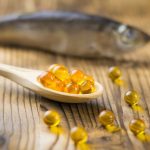 Fakanálban omega-3 kapszulák, a háttérben egy hal a faasztalon.