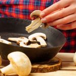 Fekete serpenyőben szeletelt gomba, melyekből az egyiket kezében tartja egy nő kockás ingben, körülötte az asztalon gombafejek.