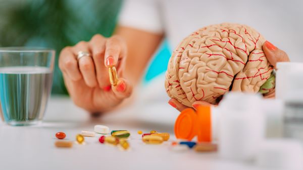 Közeli fotó doktornő kezeiről, az egyikben egy kapszulát, a másikban egy emberi agymodellt tart, mellette egy pohár víz és színes kapszulák az asztalon.