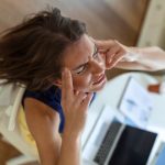 Stresszes nő az asztalnál ülve hátrahajtja fejét, előtte laptop.