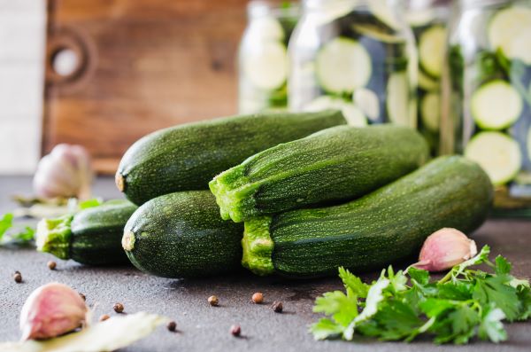 Konyhapulton egy halom cukkini, mellette fokhagymagerezdek, petrezselyemlevél, háttérben nagy befőttesüvegek cukkinikarikákkal.