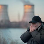 Férfi kabátban és sapkában állva orra és szája elé tartja a kezét, háttérben füstölgő gyárkémények.