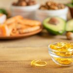 Asztalon kis üvegtálban omega-3 kapszulák, háttérben avokádó, diófélék, lazac.