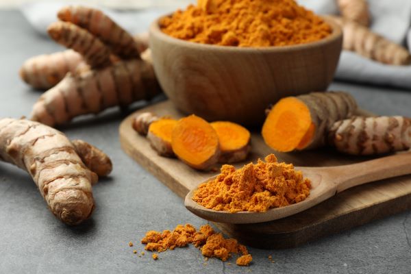 Vágódeszkán fatálban és fakanálban őrölt kurkuma, mellette kurkumagyökerek.
