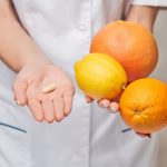 Fehér köpenyes nő egyik kezében citrusféléket tart, másikban egy vitaminkapszulát.