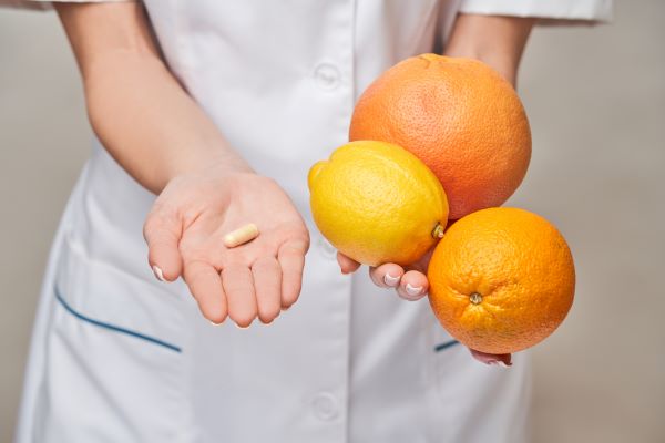 Fehér köpenyes nő egyik kezében citrusféléket tart, másikban egy vitaminkapszulát.
