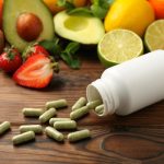 Vitaminos dobozból kidőlt kapszulák, háttérben gyümölcsök.
