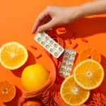 Narancssárga háttéren citrom, narancs és C-vitamin-kapszulák, amelyekért nyúl egy kéz.