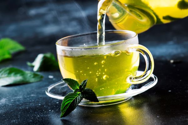 Üvegcsészébe zöld teát önt valaki üvegkancsóból.