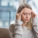 Fiatal nő fejfájással ül a kanapén, háta mögött ablak.