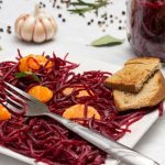 Szögletes fehér tányéron reszelt cékla mandaringerezdekkel, pirítóssal és egy villával, háttérben egy üveg cékla, egy fej fokhagyma, babérlevelek, egész borsszemek.