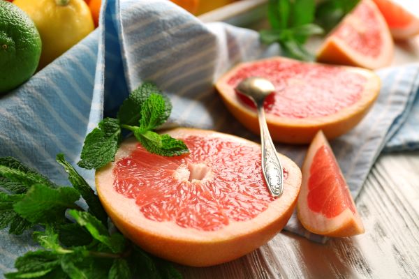 Grépfrút felszeletelve, kiskanál, kék-fehér csíkos konyharuhán, háttérben citrusfélék: citrom, lime.