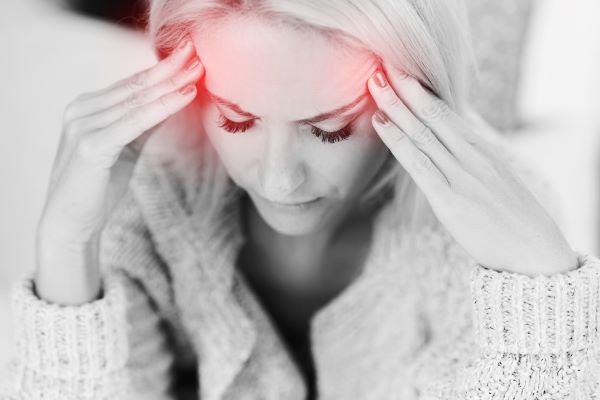 Fekete-fehér fotó nőről, aki mindkét kezével a halántékát fogja, a halántékán pirossal jelezve a fájdalom.