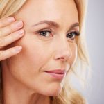Sötét karikák a szem alatt – hogyan orvosolható? Szőke nő arca közelről, egyik kezét a halántékára téve.