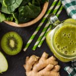 Székrekedés enyhítése almával, lenmaggal és aszalt szilvával – spenótos-gyömbéres zöld turmixital-recepttel Egy pohár zöld turmix felülnézetben, mellette egy tál spenótlevél, alma, kivi, gyömbér, zöld kockás konyharuha és 3 zöld csíkos szívószál.