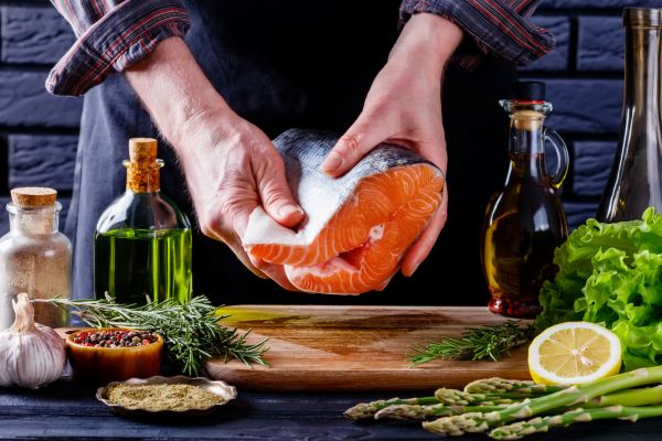 Férfi kezében tart egy darab nyers halat, előtte a konyhapulton vágódeszka, olívaolaj, citrom, rozmaring, fokhagyma, spárga.