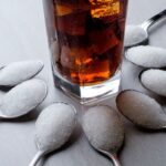 Egy pohár kóla jégkockákkal, körülötte 8 kanál, mindegyik tele cukorral.