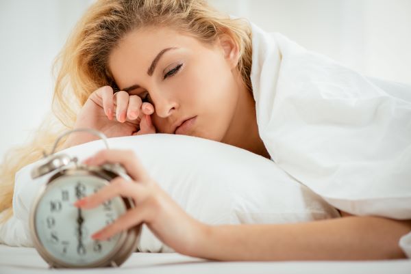 Fiatal szőke nő az ágyban álmosan nyomja ki az ébresztőórát.