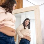 Túlsúlyos fiatal nő felsőjét felhajtva nézi magát a tükörben.