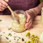 Erjesztett táplálékok, savanyú káposzta, kefir – a probiotikumok legjobb forrásai Valaki fog egy asztalon lévő befőttesüveget savanyú káposztával.