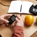 Valaki vércukormérőt tart a kezében, mellette az asztalon narancs, alma, jegyzetfüzet, toll.