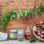 Deszkafalon zöld fűszernövények lógnak kis csokrokban, alatta egy kosárban hagyma, fokhagyma, gyömbér, fahéjrúd, csillagánizs, mellette fűszeres üvegek és mozsár.