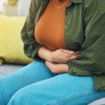 Nő a kanapén ülve mindkét kezével a hasát fogja.