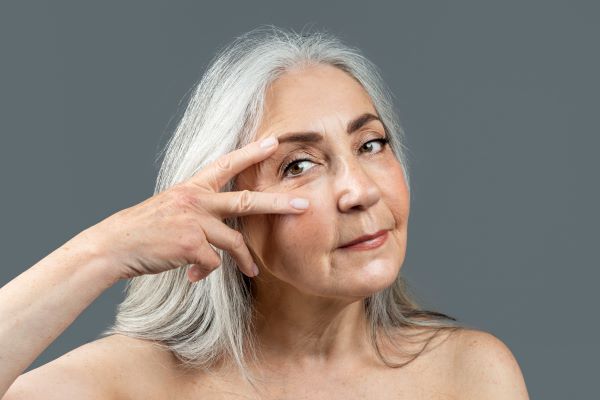 Idősebb ősz hajú nő két ujjával az arcát érinti a szeme mellett.